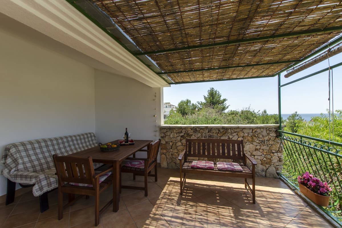 A covered outdoor seating area with a table and chairs, a bench, and a view of the sea.