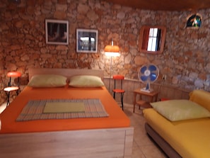 Una habitación con pared de piedra, una cama con ropa de cama naranja, un sofá amarillo, una mesita con un ventilador y dos lámparas.