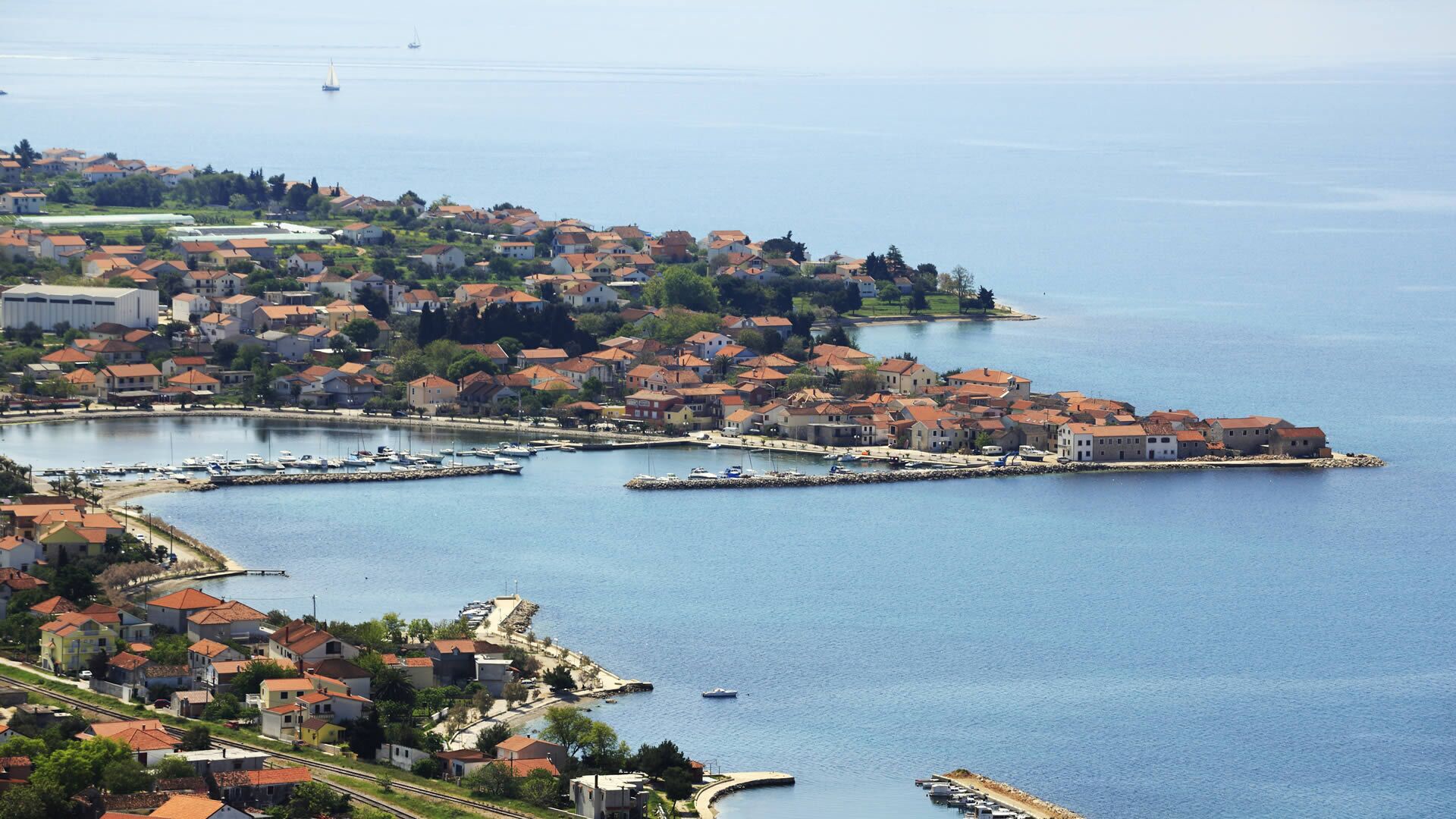 Uma vila costeira com um porto de iates, casas com telhados vermelhos e vista clara para o mar.