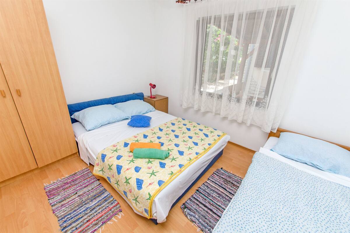 Une chambre à coucher comprenant un lit, une table de chevet, une fenêtre avec des rideaux légers et une armoire en bois.