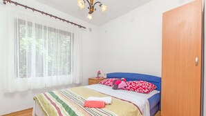 Ein Schlafzimmer mit Bett, Nachttisch, Kleiderschrank, einem Fenster mit durchsichtigen Vorhängen und einer Deckenleuchte.