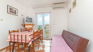 Ein kleiner, gut beleuchteter Raum mit Esstisch, Stühlen, einer Couch und einem Fernseher. Ein Balkon bietet Blick auf Gebäude.