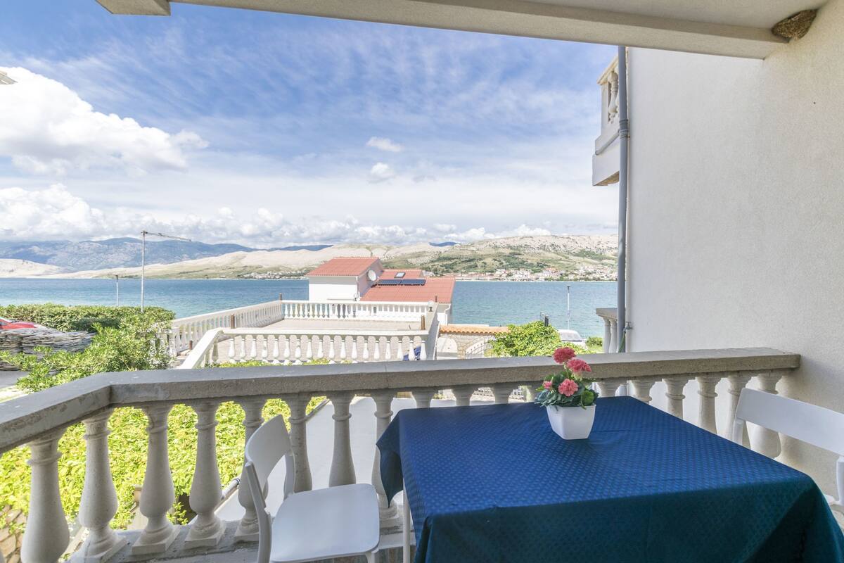 A balcony with a table and chairs, overlooking a coastal view with mountains and a body of water.