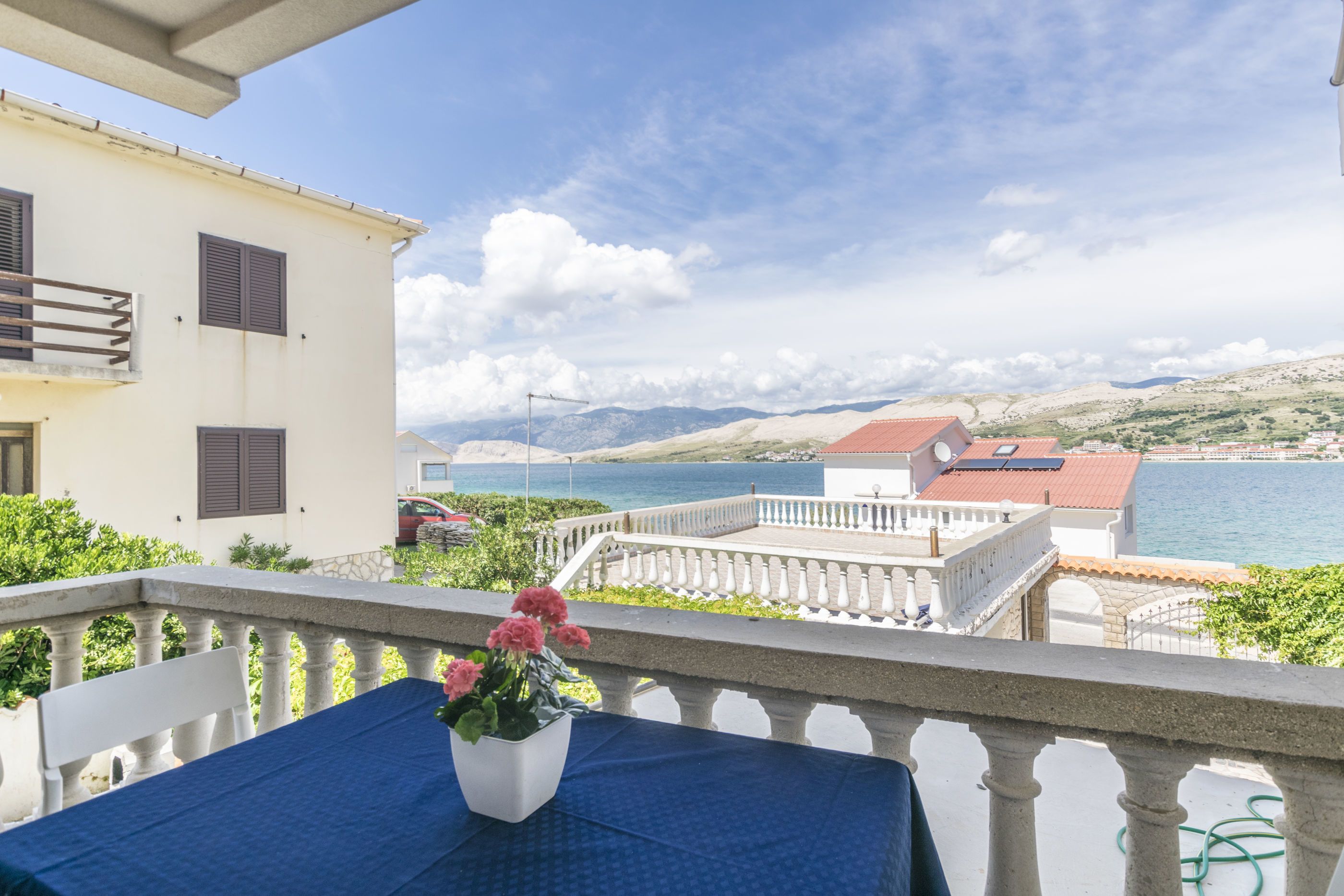 A balcony with a table and a potted plant, overlooking a coastal view with buildings and mountains.