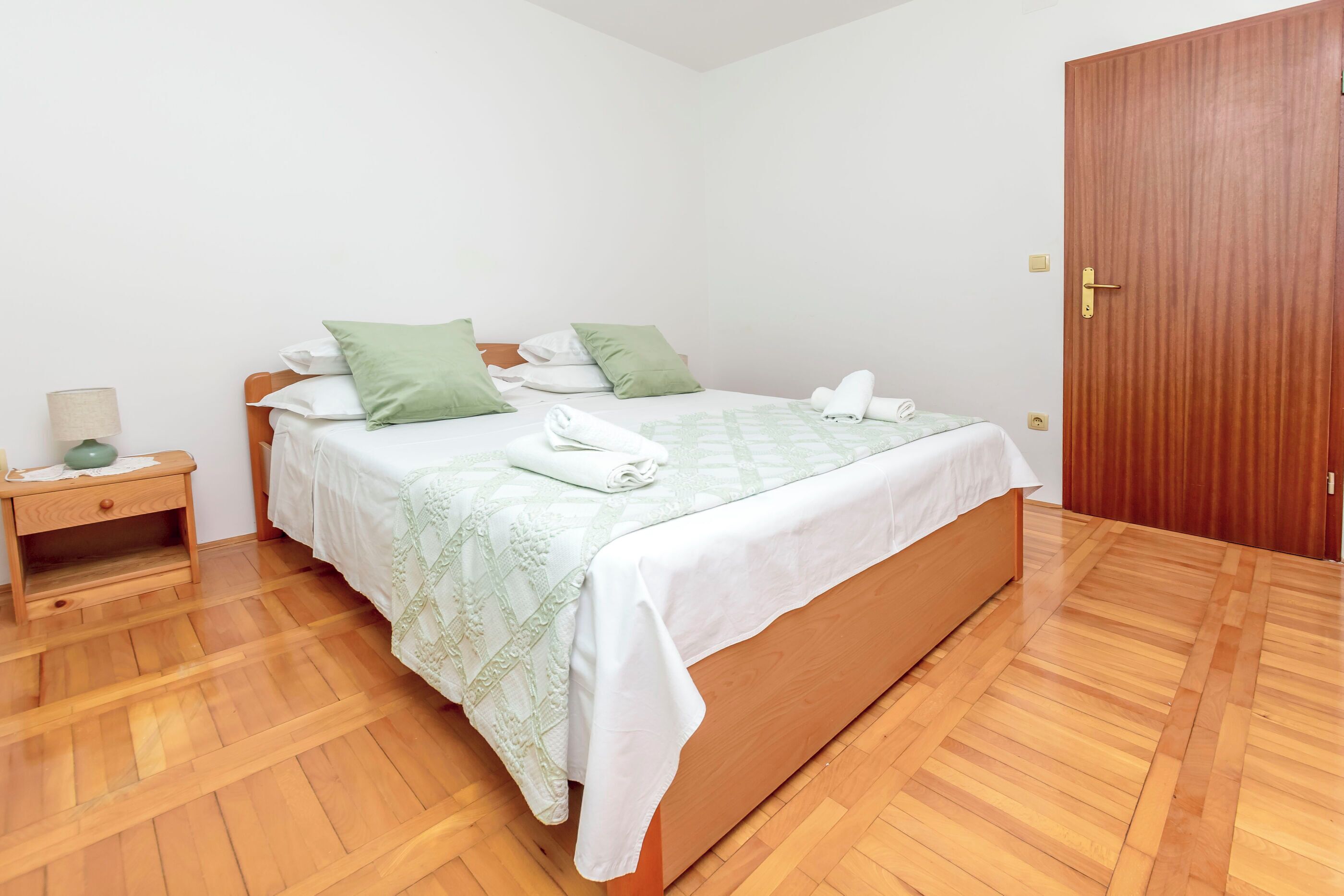 A neatly made bed with a green and white bedspread, a wooden nightstand with a lamp, and a wooden door.