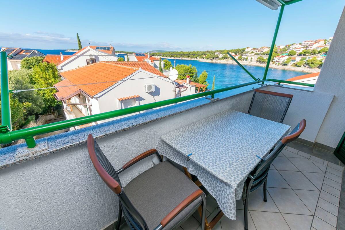 Ein Balkon mit Tisch und Stühlen, mit Blick auf eine Küstenstadt mit Häusern und blauem Meer.
