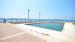 A swimming pool with steps leading into the water, surrounded by a paved area with a clear view of the sea and distant hills. - Apartment Slavko (Biograd)