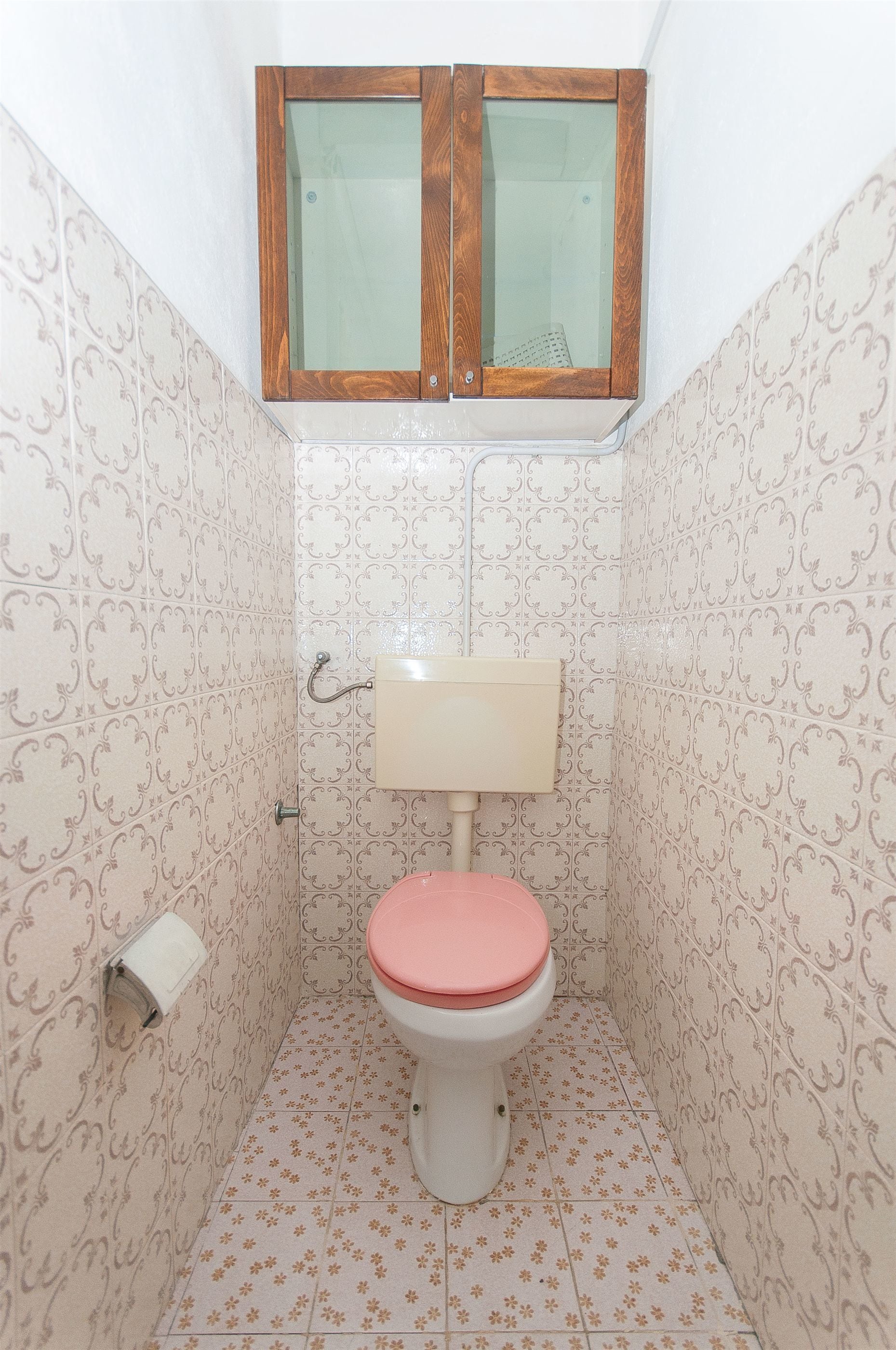 Ein kleines Badezimmer mit Toilette, einem Holzschrank darüber, strukturierten Tapeten und einem Toilettenpapierhalter.