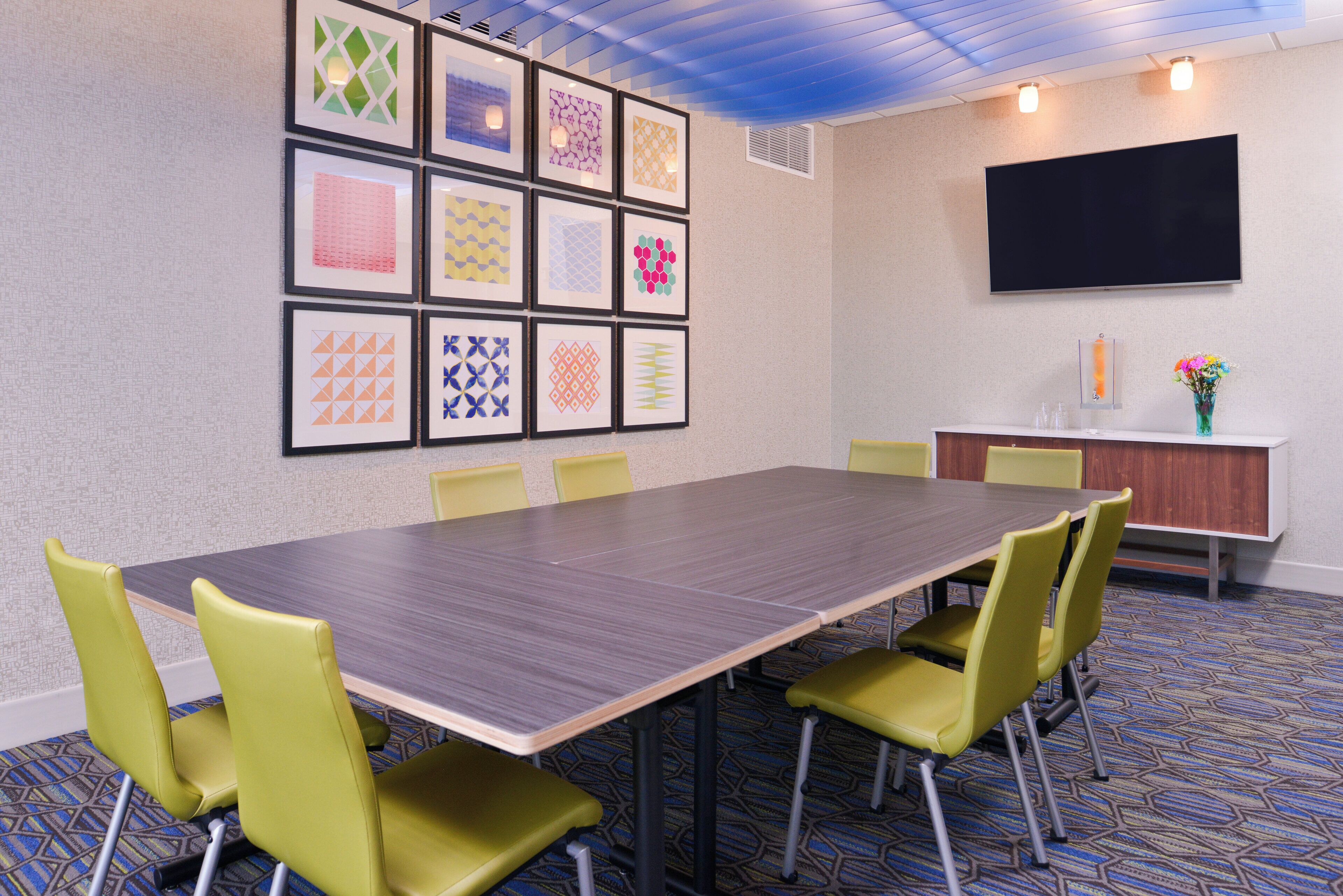 A conference room with a long table, six chairs, a flat-screen TV, a small cabinet, and framed artwork on the wall.