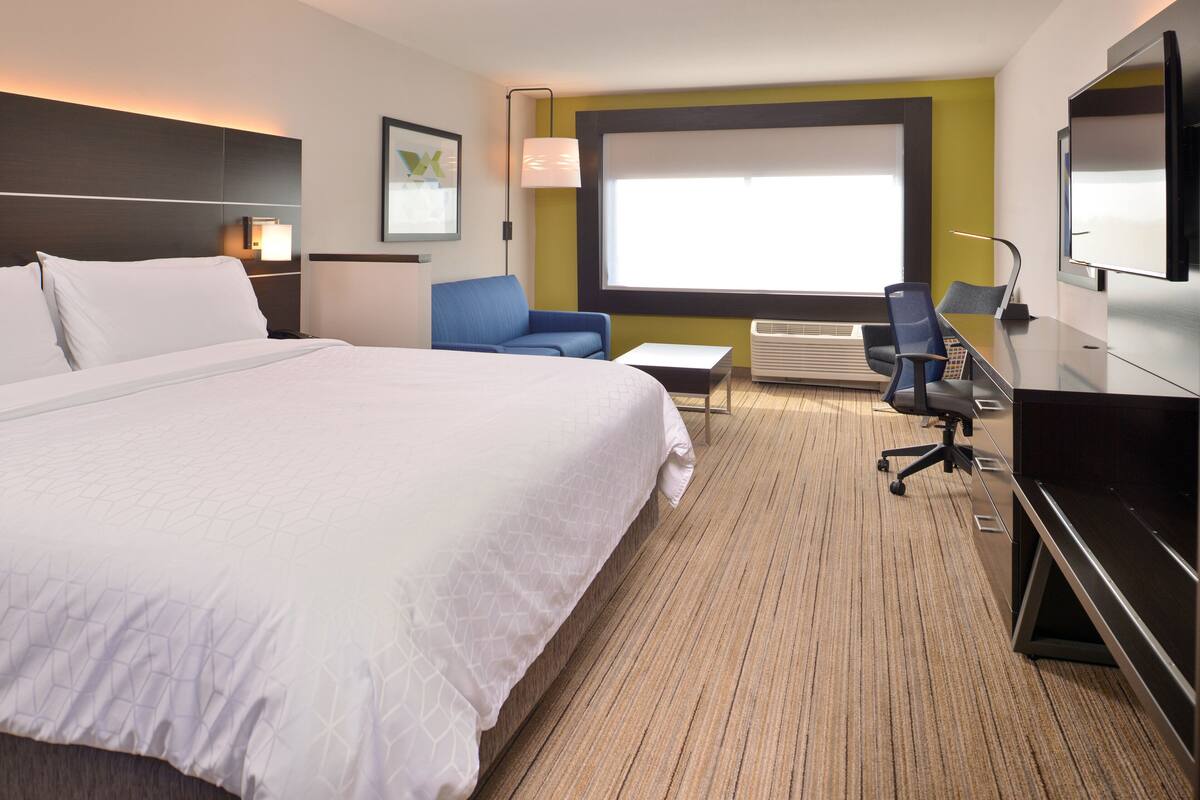 A hotel room with a large bed, a desk, two chairs, a TV, and a window with blinds.