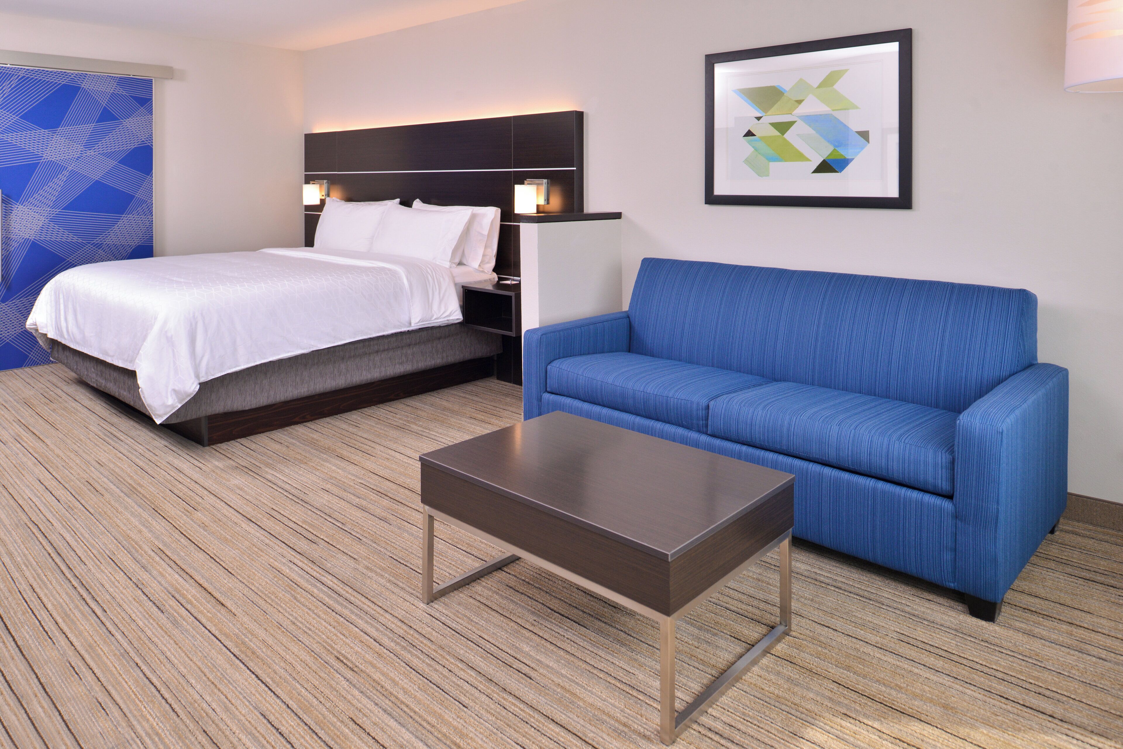 Ein Hotelzimmer mit einem Bett, einem blauen Sofa, einem kleinen Tisch und einer Wand mit einem geometrischen Kunstwerk.