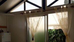 A room with a ceiling fan, a refrigerator, a table with a vase of flowers, and a view of greenery through a window with sheer curtains.