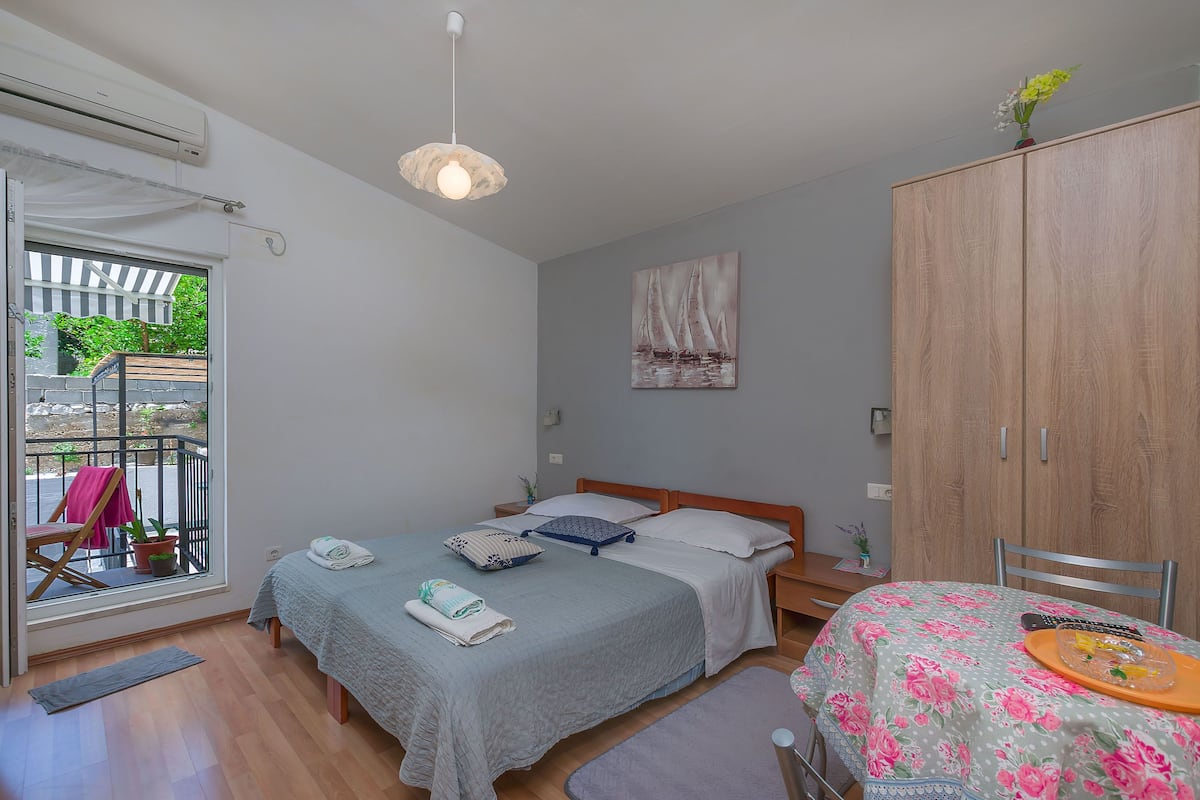A bedroom with a bed, a wooden wardrobe, a dining table with a floral tablecloth, and a window with a view of greenery.