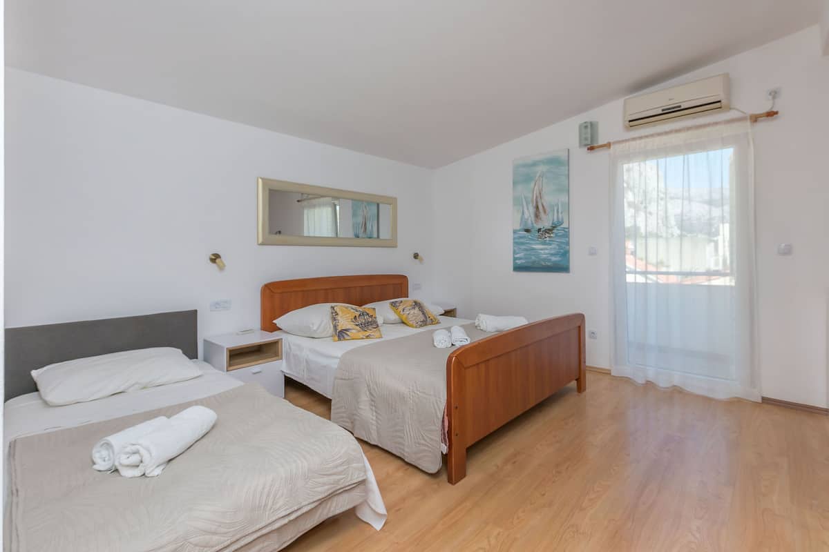 A hotel room with two beds, a wooden floor, a painting of a sailboat, an air conditioning unit, and a window with a curtain.