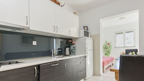 A modern kitchen with dark cabinetry, a white countertop, a microwave, and a refrigerator. There is a window with a view of trees and a bed with a red blanket in the adjacent room. - Apartments Glorija (Rogoznica)