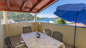 A balcony with a table and chairs, a blue umbrella, and a view of a lake and hills. - Apartments Glorija (Rogoznica)