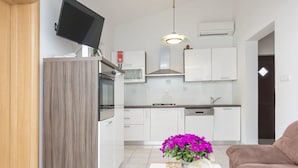 A modern kitchen with white cabinets, a built-in oven, and a microwave. There is a brown couch and a table with a vase of purple flowers. - Apartments Glorija (Rogoznica)