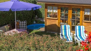 En træpavillon med swimmingpool, omgivet af frodig vegetation og en terrasse med bord og stole.