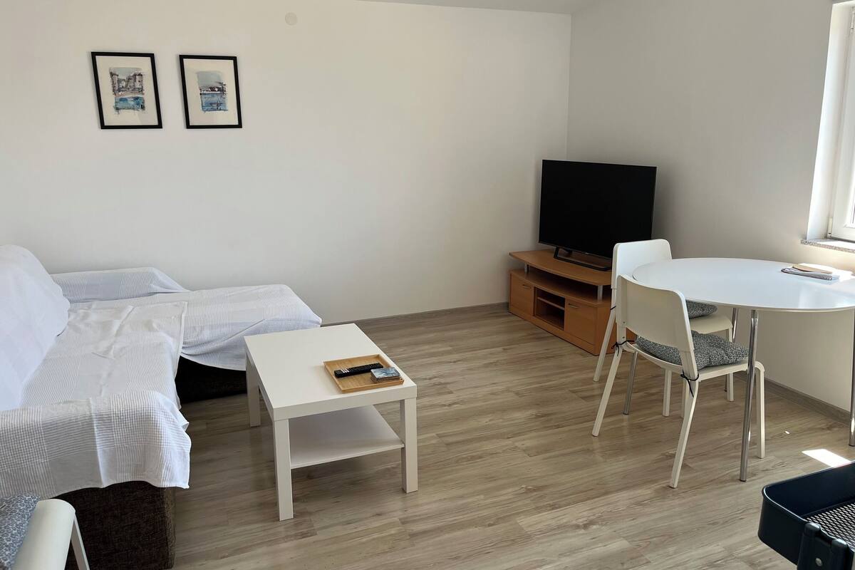 Una sala de estar moderna con un sofá, una pequeña mesa blanca, un televisor sobre un soporte de madera y un comedor con una mesa y sillas blancas.