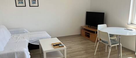 Ein modernes Wohnzimmer mit einer Couch, einem kleinen weißen Tisch, einem Fernseher auf einem Holzgestell und einem Essbereich mit weißem Tisch und Stühlen.