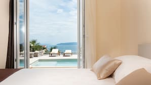 Ein Hotelzimmer mit Bett, Blick auf Pool und Liegestühle, und blauem Himmel.