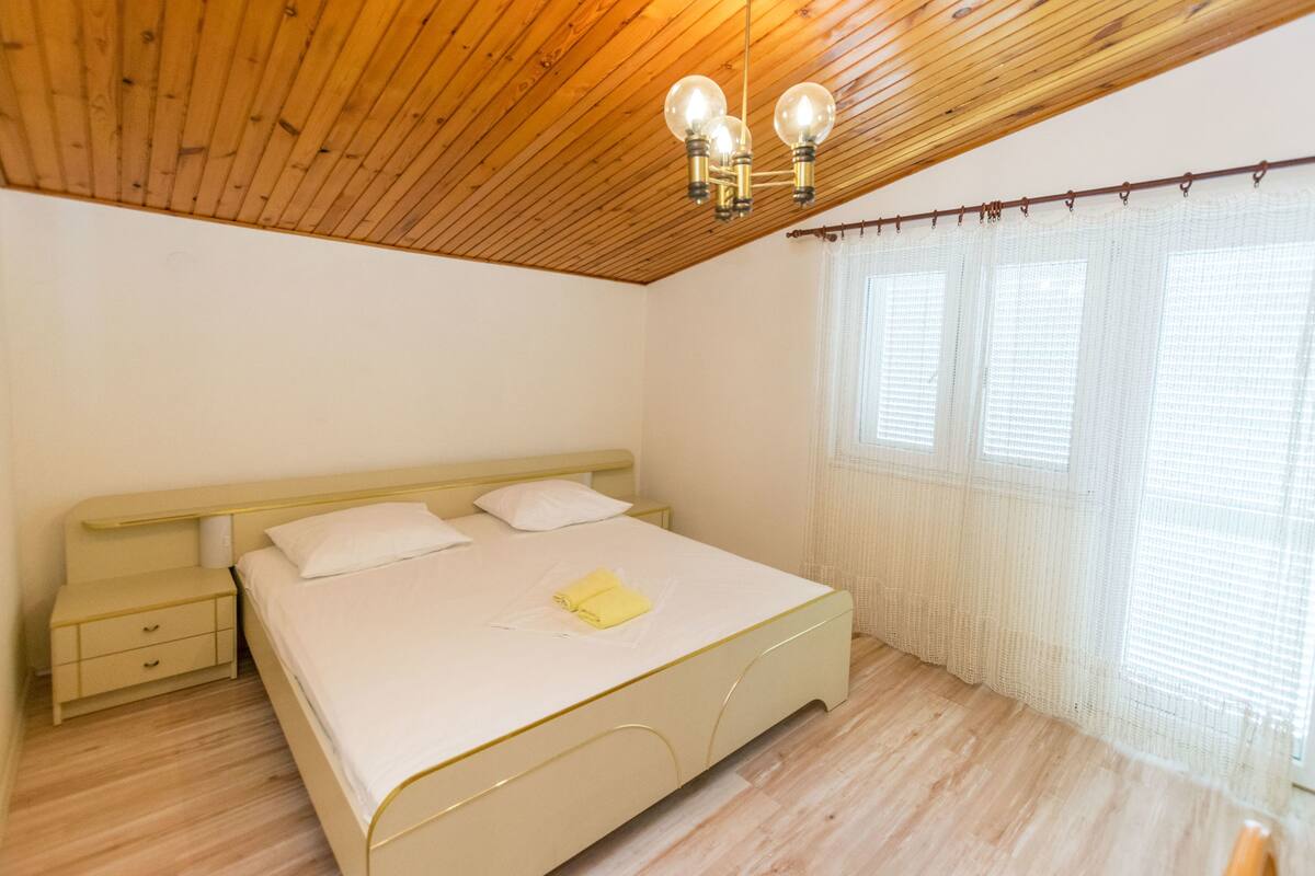 A bedroom with a wooden ceiling, a bed with white bedding, a nightstand, and a window with blinds.