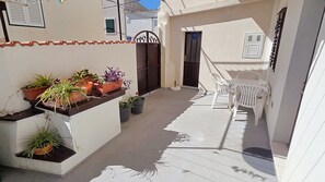 Un estrecho pasillo exterior con macetas, una mesa y sillas blancas, y una puerta con reja de metal.