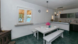 Un coin repas avec une table et des bancs blancs, une fenêtre donnant sur un beau paysage et une cuisine avec des meubles sombres et un évier.