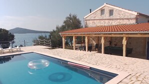 Une piscine avec une terrasse en pierre, un espace extérieur couvert avec des sièges, et une vue sur la mer.