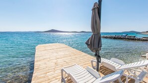 Une jetée en bois s’étendant dans une eau d’un bleu limpide, bordée d’une rangée de chaises longues blanches et surmontée d’un parasol gris.