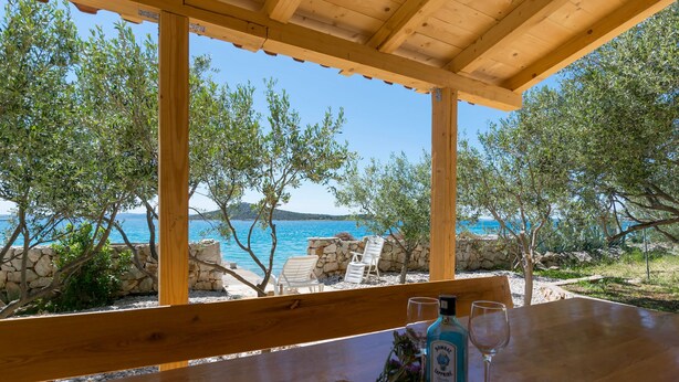 Une table en bois, sur laquelle se trouvent une bouteille de spiritueux et un verre, donnant sur une plage bordée de murs de pierre et de chaises blanches.