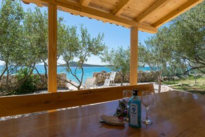 A wooden table with a bottle of liquor and a glass, overlooking a beach with stone walls and white chairs. - House Hacienda Stoerman (Tkon)