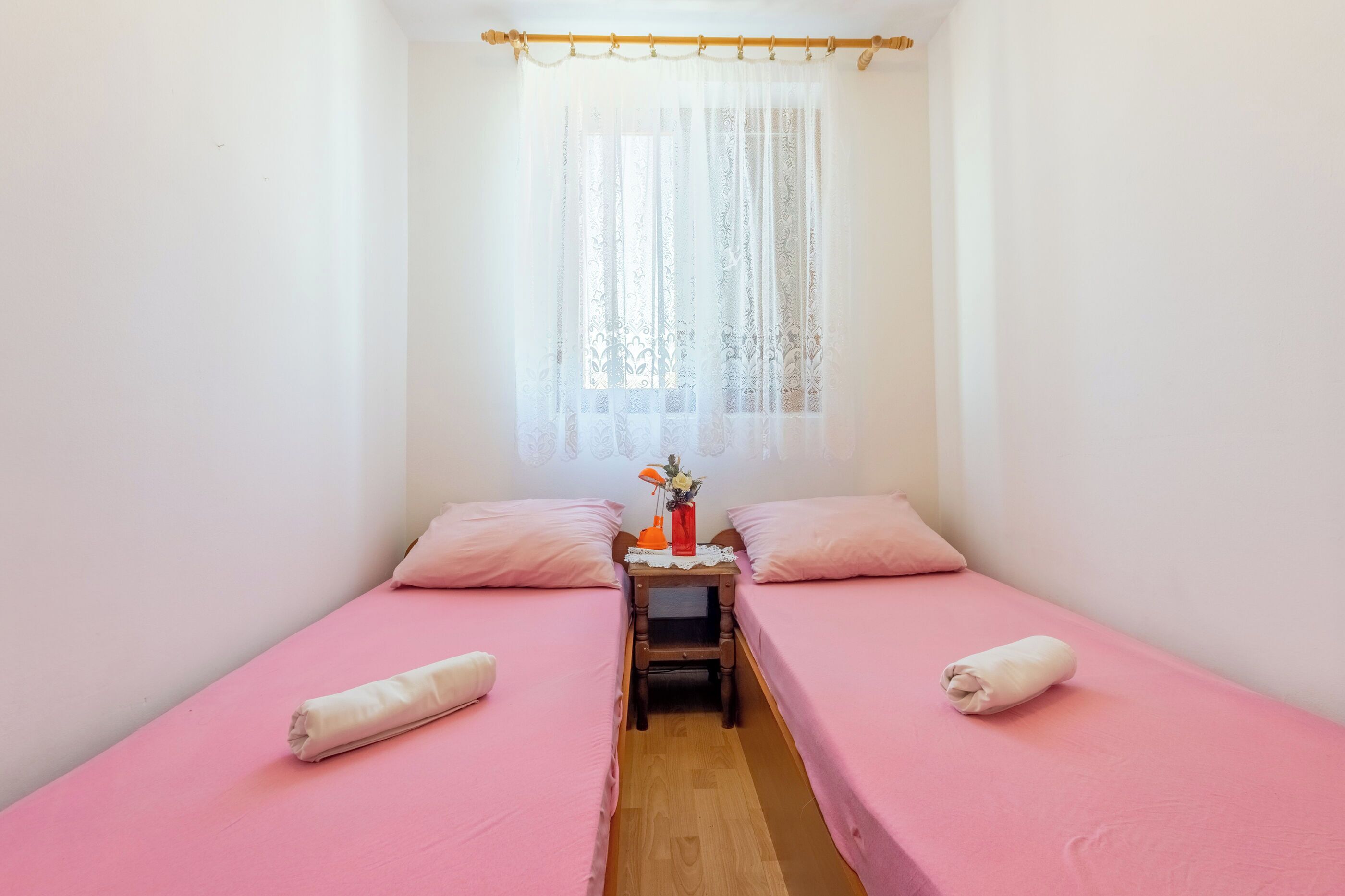 Une petite chambre propre, avec deux lits, une petite table sur laquelle se trouvent un vase et une lampe, et une fenêtre ornée de rideaux légers.