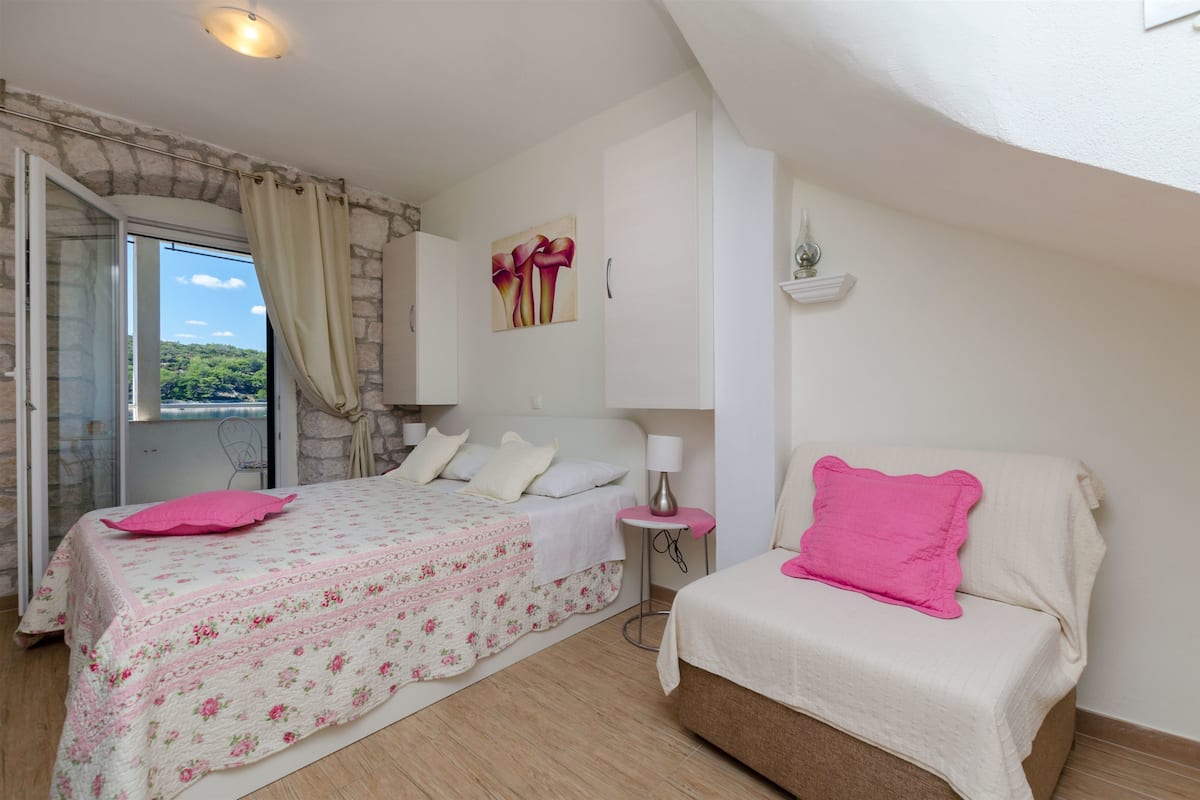 A bedroom with a bed, a bedside table, a lamp, and a view of greenery outside.