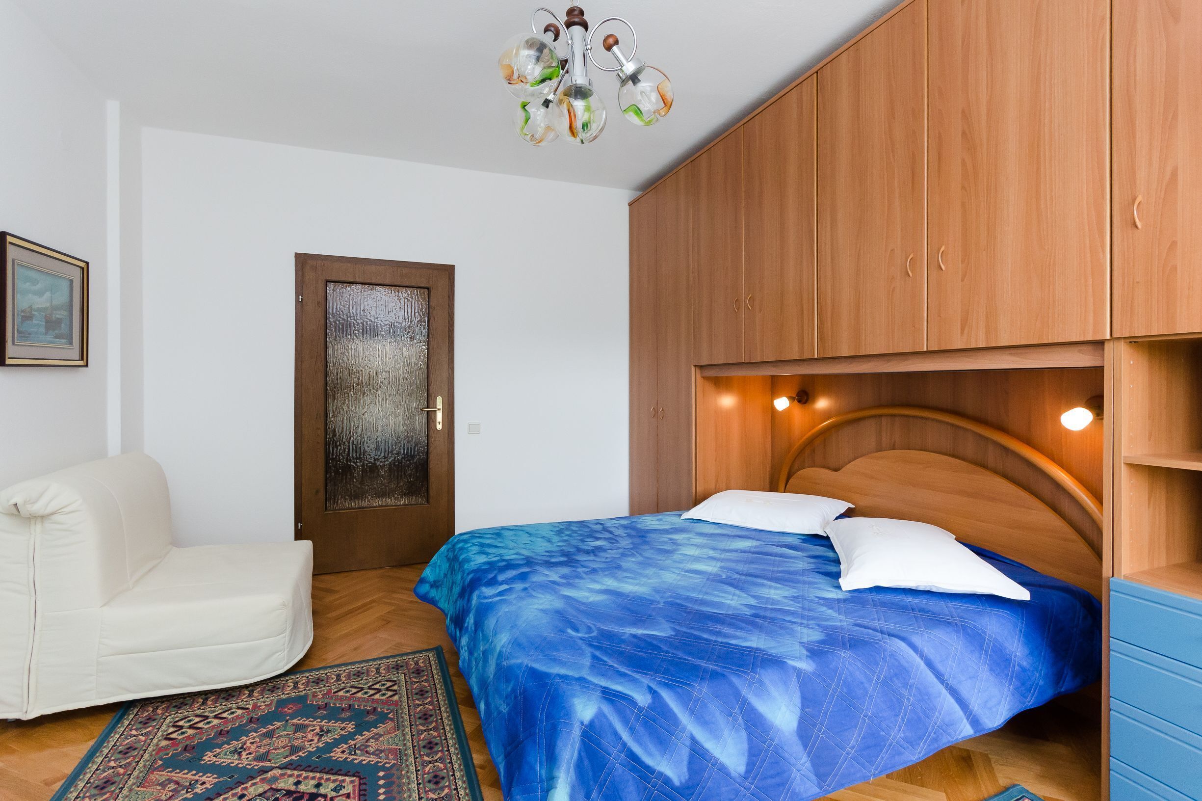 A bedroom with a bed, wooden wardrobe, a white chair, and a patterned rug.