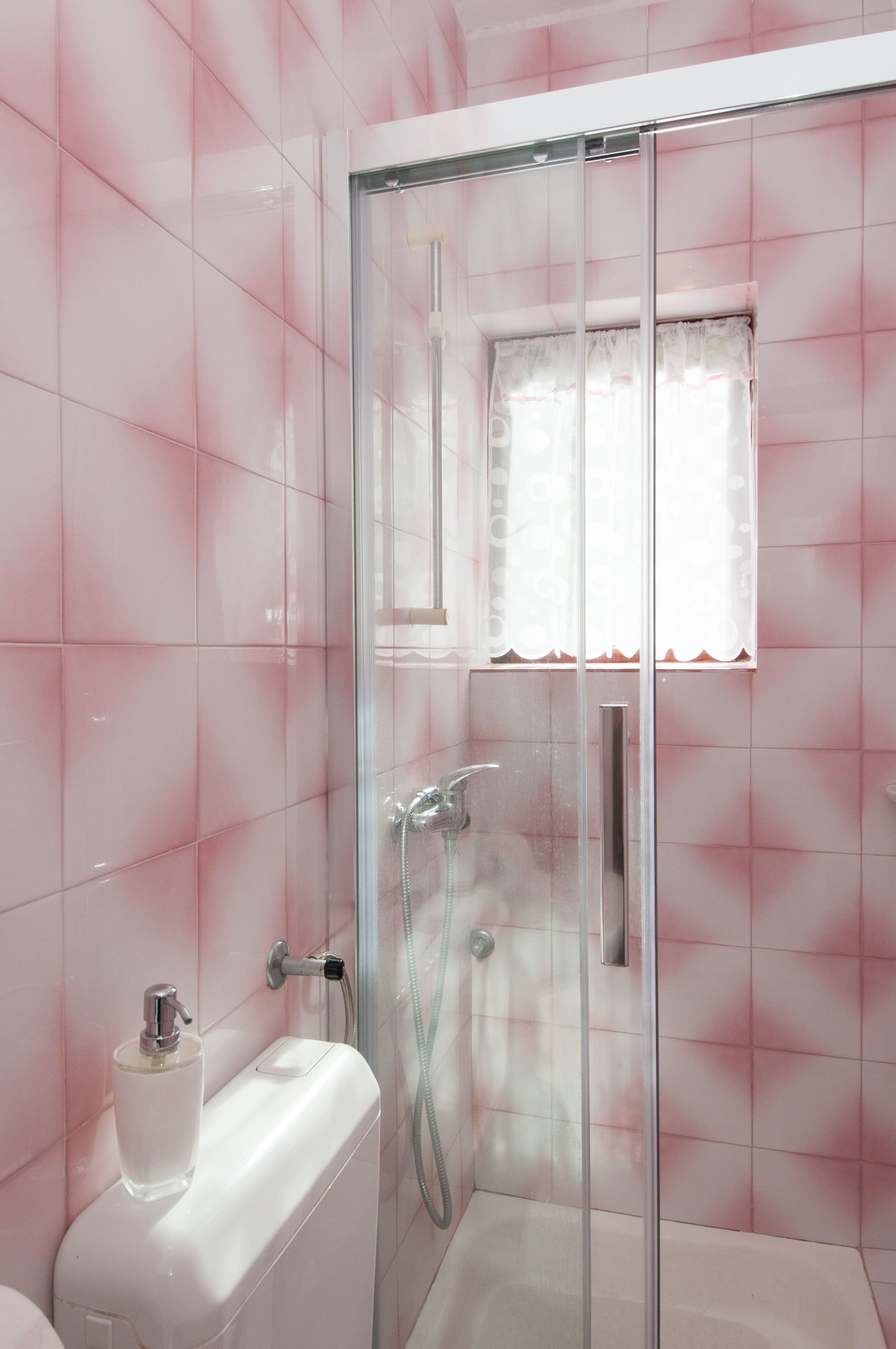 A bathroom with a glass-enclosed shower, pink tiled walls, a toilet, and a window with a white curtain.