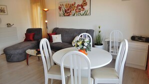 Uma sala de estar com um sofá cinzento, uma mesa de jantar branca com quatro cadeiras, uma planta em vaso e um quadro na parede.
