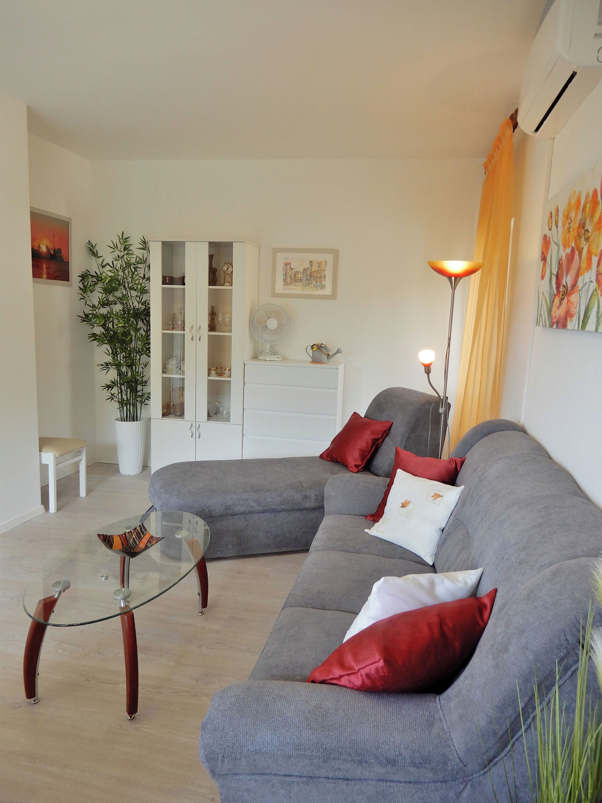 Una sala de estar moderna con un sofá seccional gris, una mesa de centro de vidrio, un mueble blanco, una lámpara de pie y una planta grande en maceta.