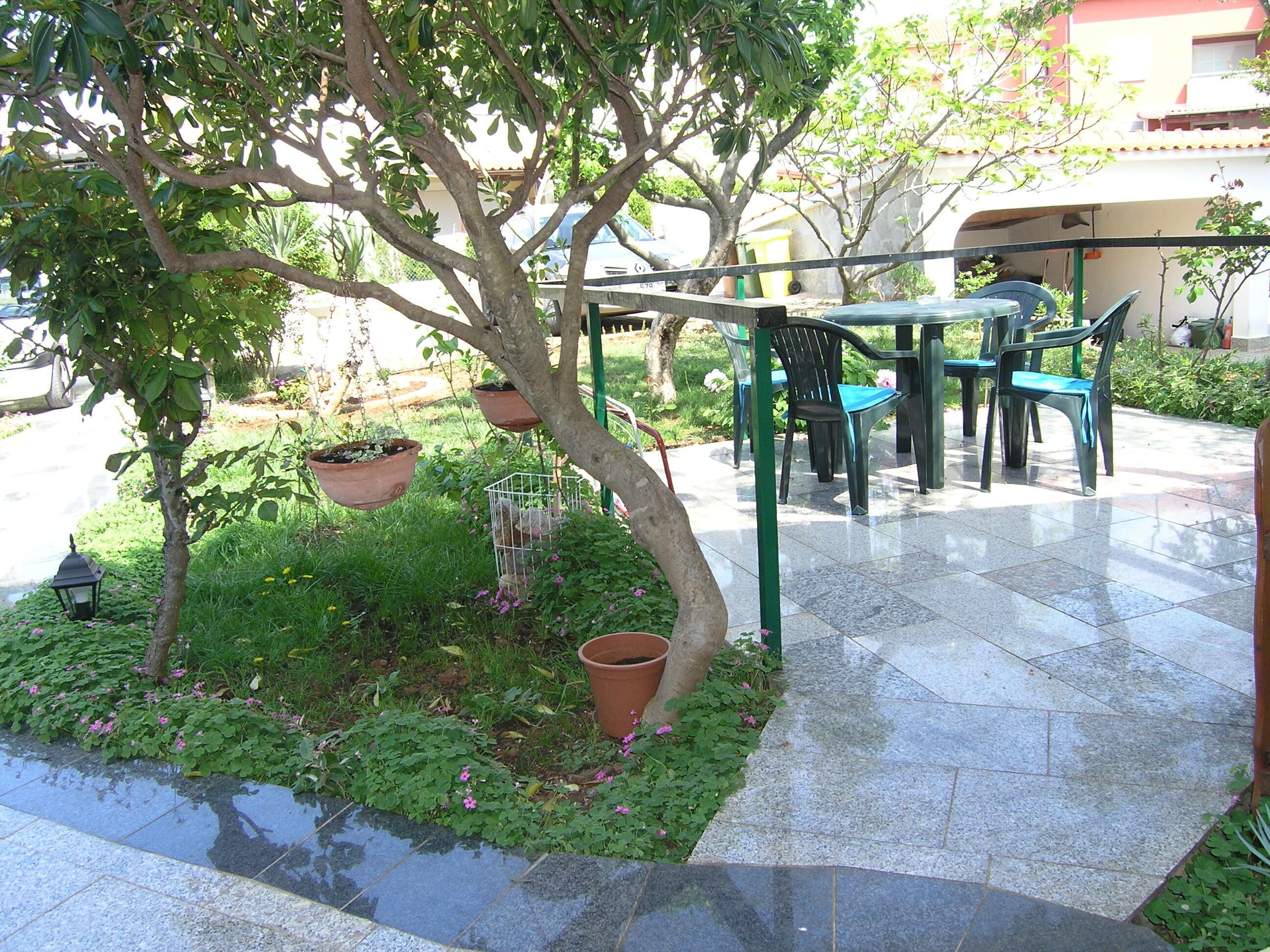An outdoor seating area with a table and chairs, surrounded by potted plants and a tree.