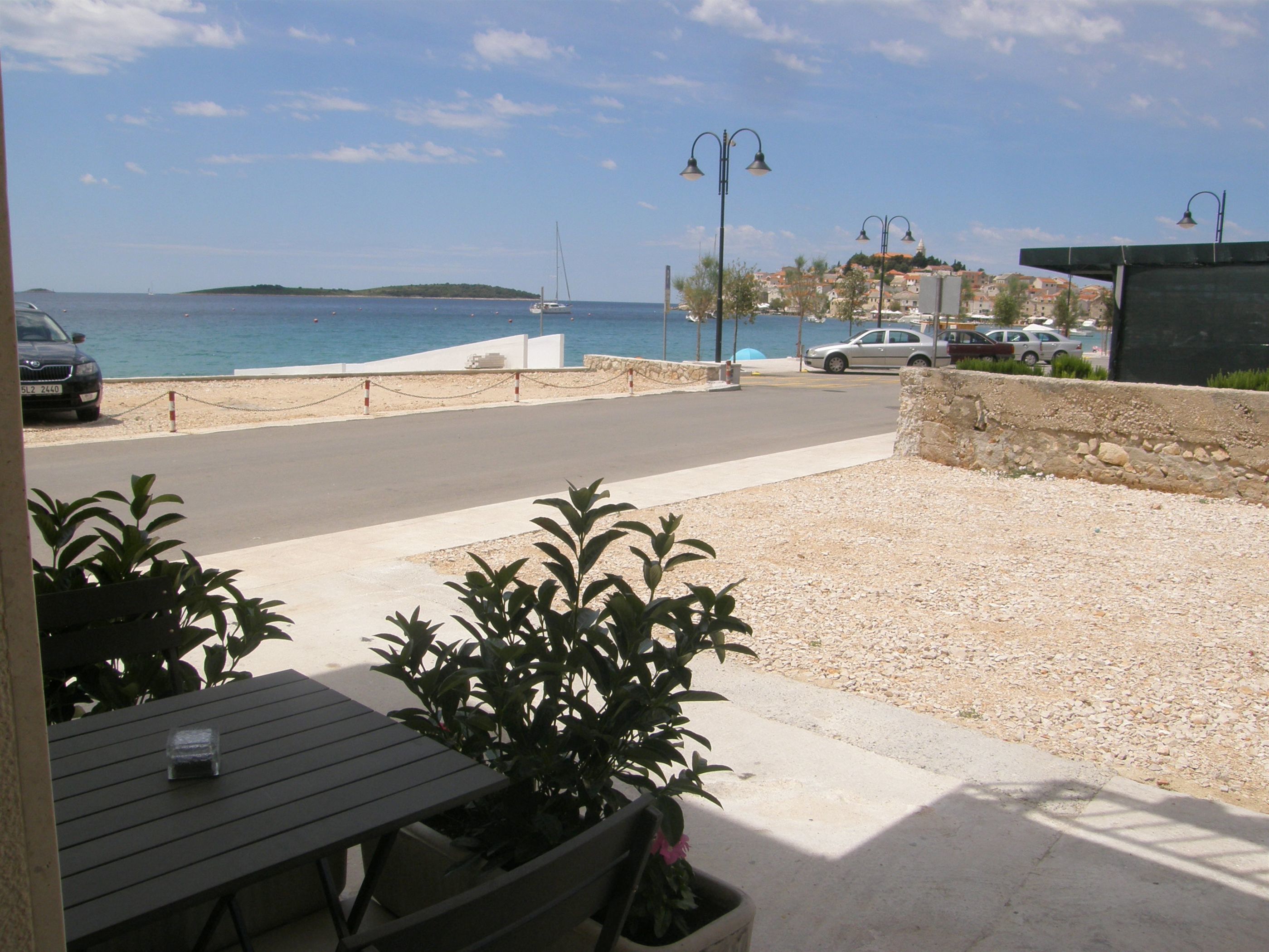 A coastal view with a road leading to the sea, a parked car, and a building with a balcony overlooking the water.
