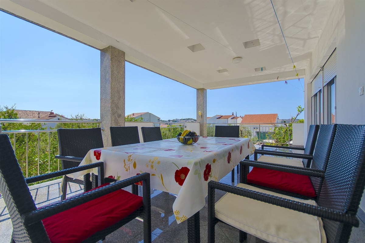 A covered outdoor dining area with a table set for four, surrounded by wicker chairs with red cushions.