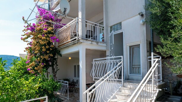 Een gebouw met twee verdiepingen, een balkon, stenen trappen en een tuin met paarse bloemen.