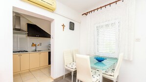 Een compacte keuken met een gasfornuis, een spoelbak en een raam met zonwering. Er staat een eettafel met stoelen bij, en een blauwe kom erop.