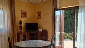 A dining area with a round table, chairs, a chandelier, a television, and a view through a glass door to a garden. - Apartments Stana (Zadar)