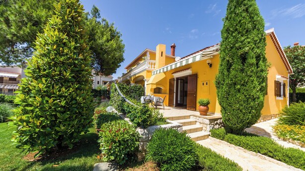 A yellow building with a stone staircase leading to a door, surrounded by greenery and a clear blue sky. - Apartments Stana (Zadar)