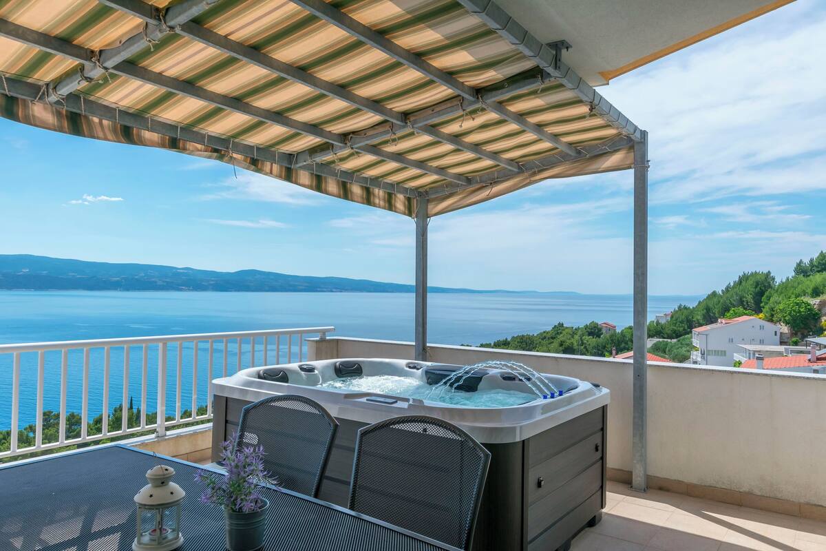 A balcony with a hot tub, a table with chairs, and a view of the sea.