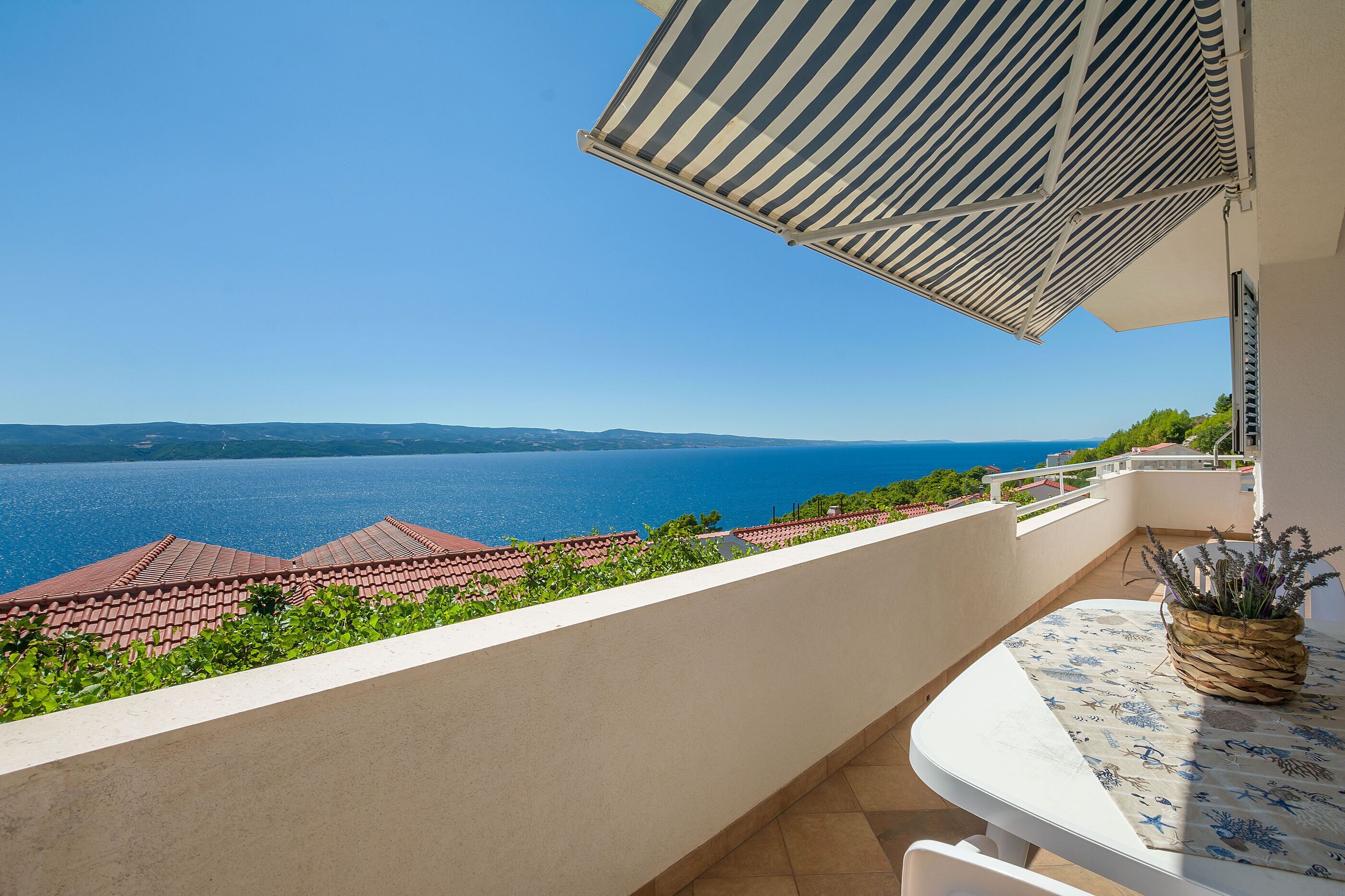 Un balcon avec vue sur la mer, une table agrémentée d’une plante et un auvent à rayures.