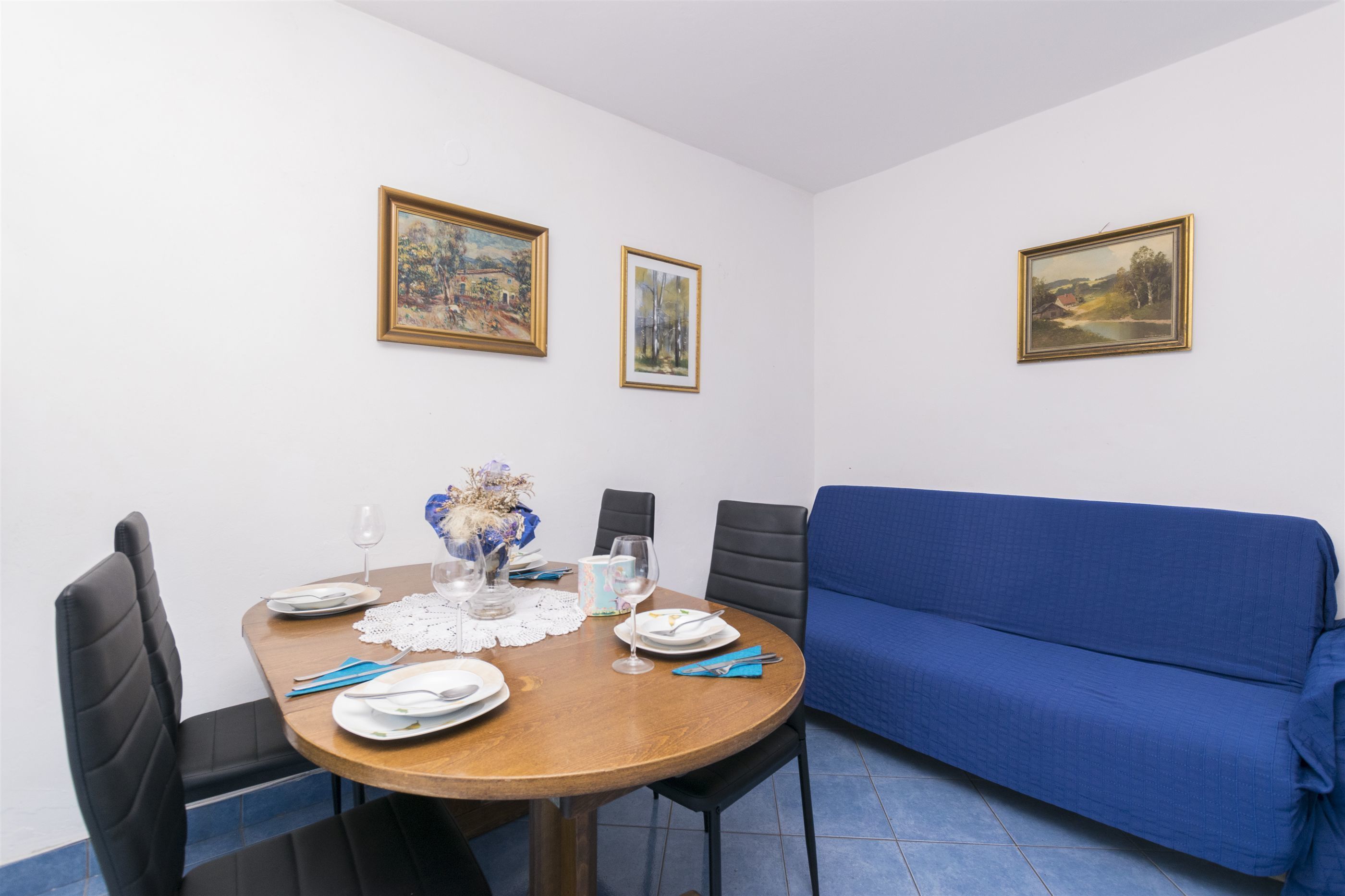 A dining area with a wooden table set for four, a blue sofa, and three framed paintings on the wall.