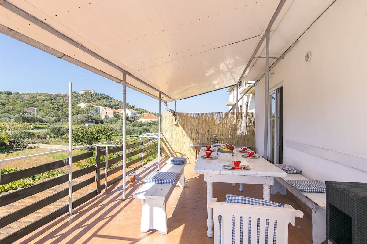 Un área de comedor al aire libre techada, con mesas blancas y cojines blancos y azules a cuadros, con vista a una colina con edificios.