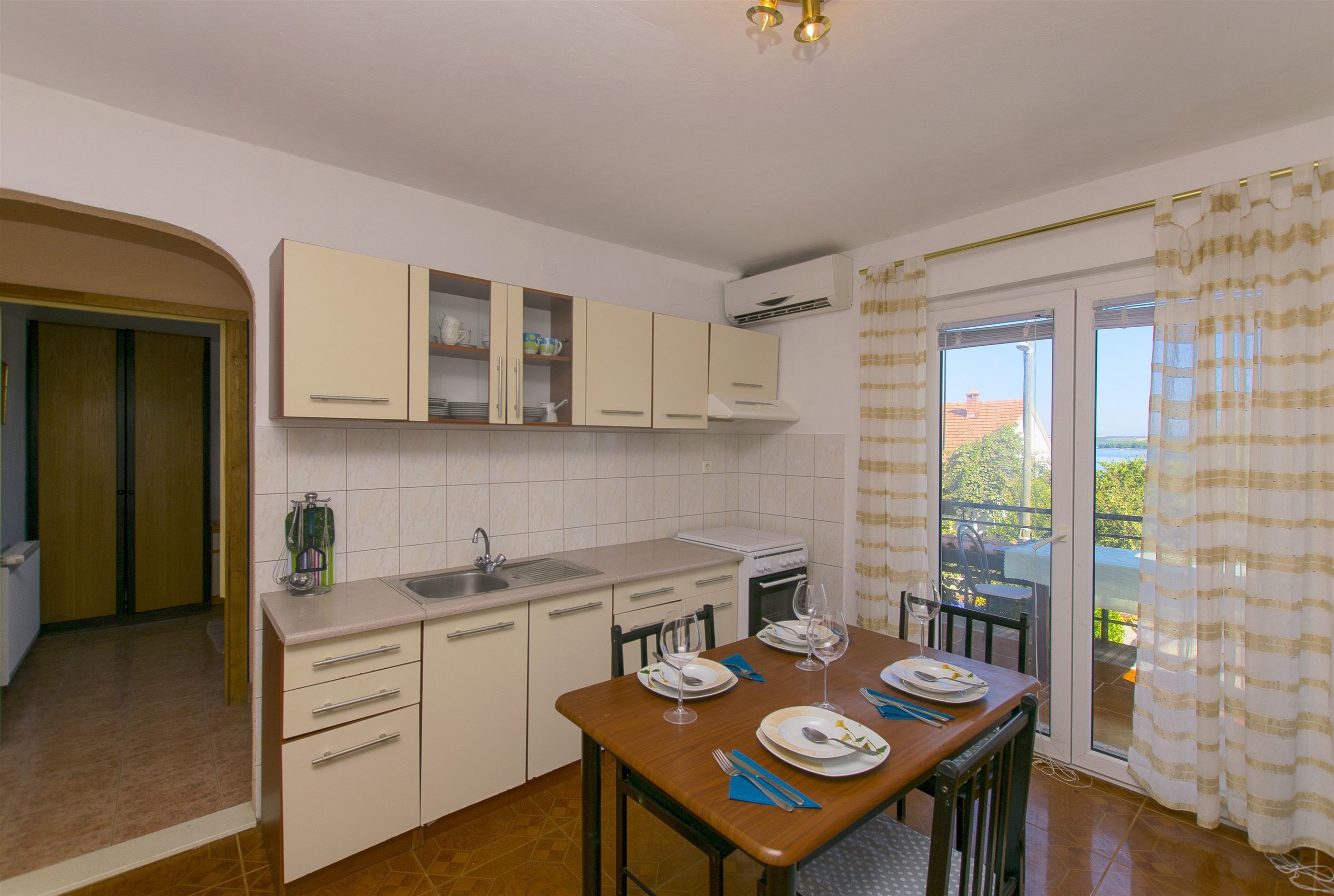 Une cuisine avec des meubles blancs, une table à manger dressée avec des assiettes et des couverts, et une vue sur l’extérieur à travers la porte vitrée.